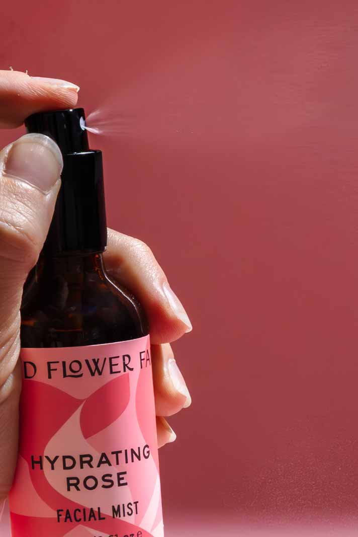 A hand sprays a bottle from the good-flower-farm Seasonal Skincare Bundle against a pink background, with a visible mist coming out of the nozzle.