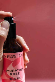 A hand sprays a bottle from the good-flower-farm Seasonal Skincare Bundle against a pink background, with a visible mist coming out of the nozzle.