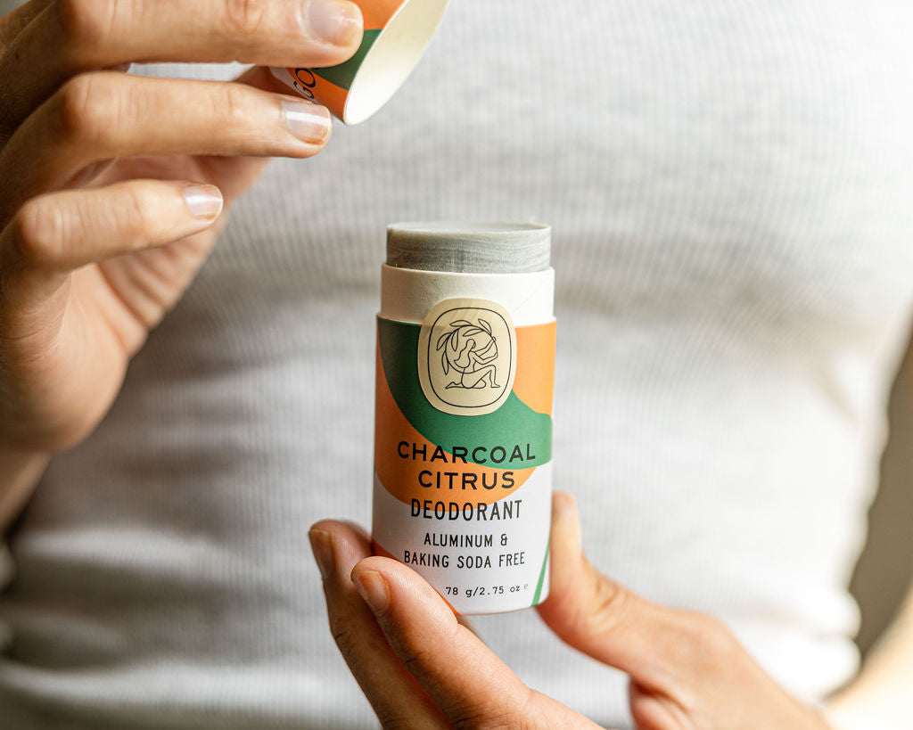 A person in a white tank top holds a Good Flower Farm Charcoal Citrus Deodorant Plastic-Free Tube, labeled aluminum & baking soda free. The biodegradable tubes cap is being removed with one hand while held with the other.