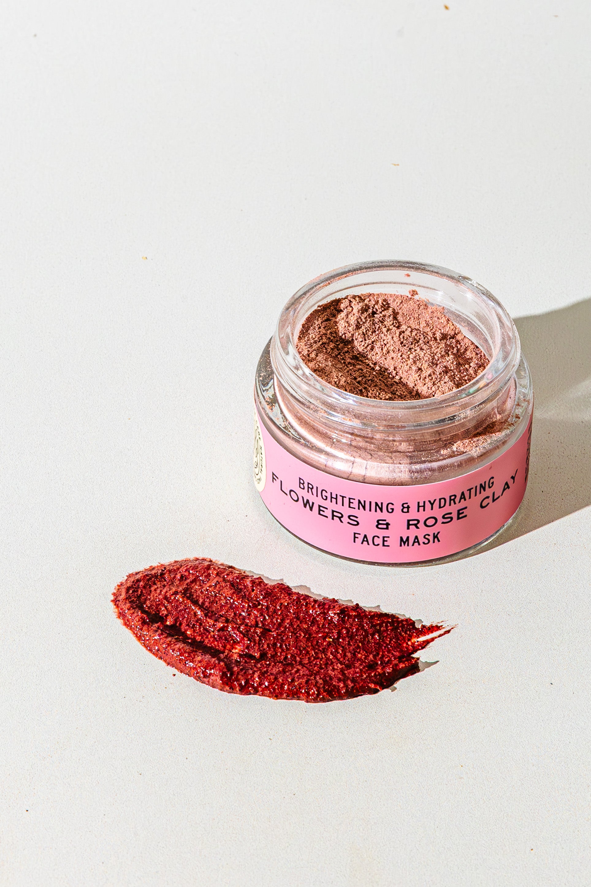 An open jar of TESTER — Flowers & Rose Clay Botanical Face Mask by Good Flower Farm rests on a light surface, showcasing its botanical formula with a swatch of the rosy clay mask displayed in front.