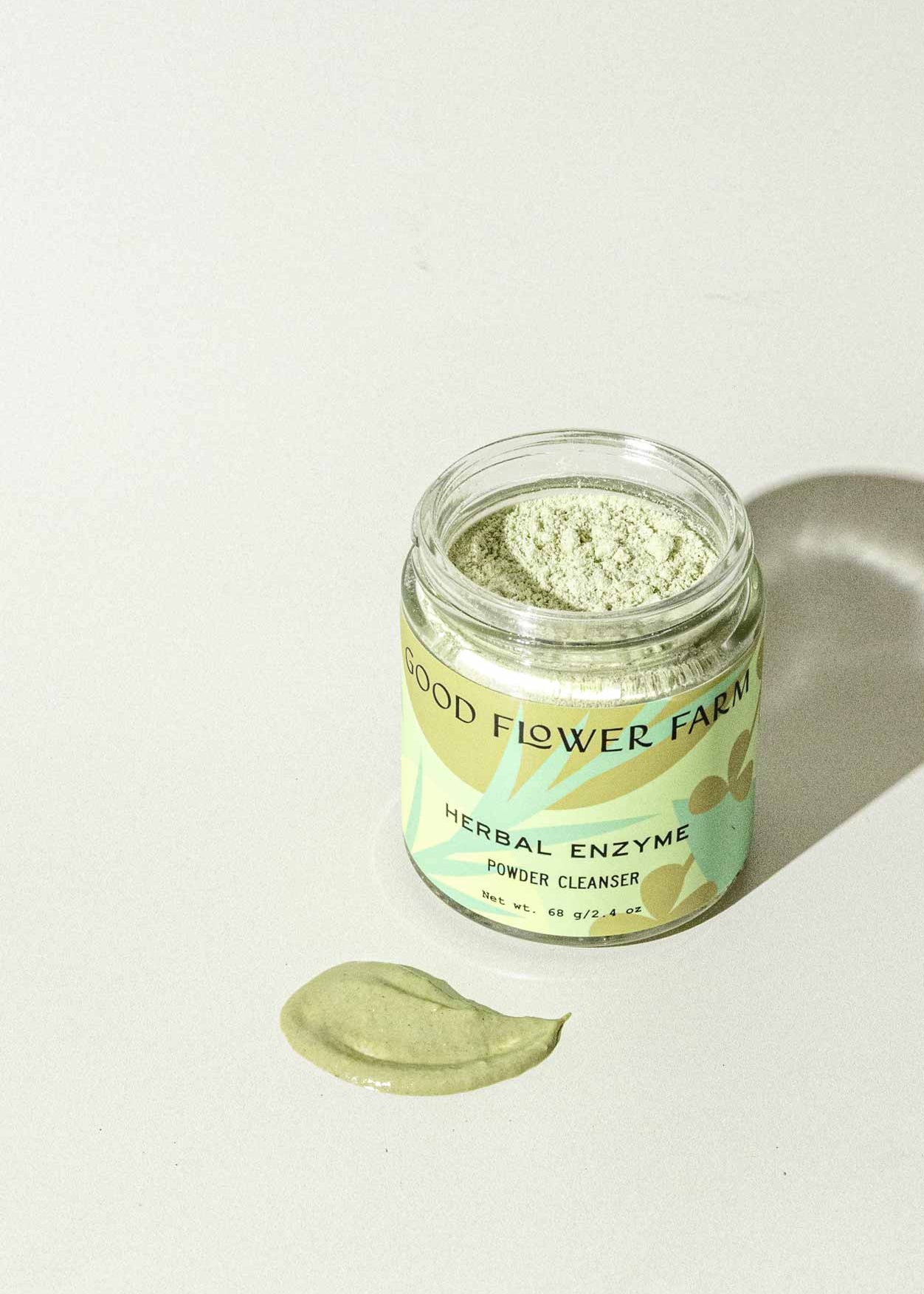 A jar of Good Flower Farm’s TESTER — Herbal Enzyme Powder Facial Cleanser rests on a light surface with green powder, formulated with coconut milk and aloe vera, displayed in front. The label features a pale green background with yellow and brown designs.