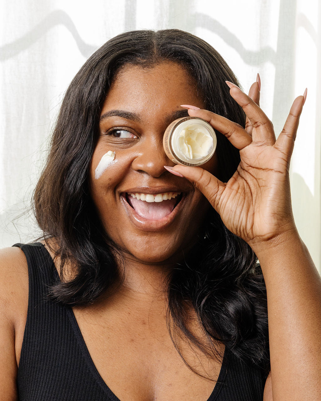 A smiling woman in a black tank top holds up a small jar of good-flower-farm’s Dry Skin Bundle: Save 15% to her eye, with a dab of cream on her cheek. She has long hair and stands in front of a white curtain, showcasing the importance of facial moisturizing for dry skin.