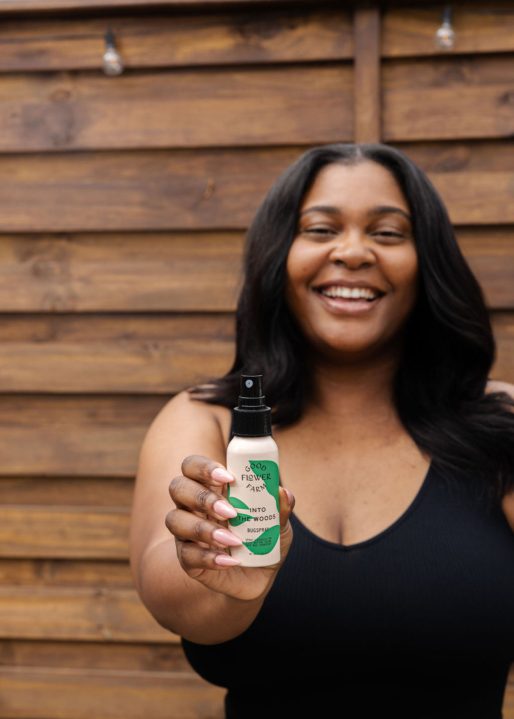 A smiling woman in a black tank top holds the good-flower-farm Seasonal Skincare Bundle toward the camera, standing in front of a wooden fence.