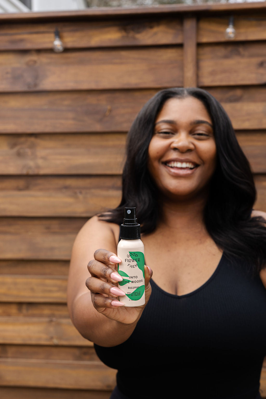 A smiling woman in a black tank top holds the good-flower-farm Seasonal Skincare Bundle toward the camera, standing in front of a wooden fence.