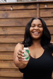 A smiling woman in a black tank top holds the good-flower-farm Seasonal Skincare Bundle toward the camera, standing in front of a wooden fence.
