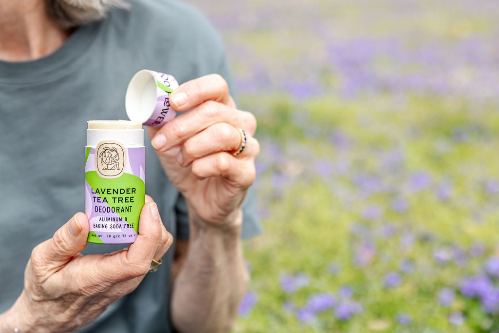 Someone in a gray shirt holds an open Good Flower Farm Lavender Tea Tree Deodorant Plastic-Free Tube, with a blurred field of purple flowers in the background.