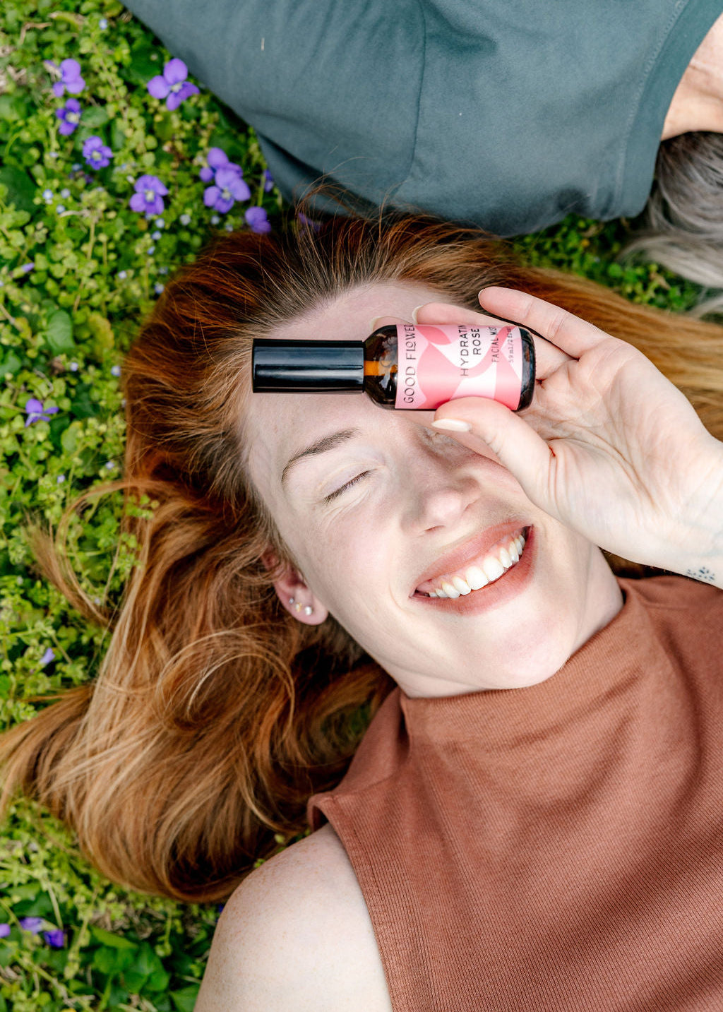 A smiling woman lies on grass and purple flowers, holding the good-flower-farm Seasonal Skincare Bundle’s pink spray bottle over one eye while wearing a sleeveless brown top.