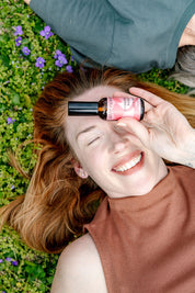 A smiling woman lies on grass and purple flowers, holding the good-flower-farm Seasonal Skincare Bundle’s pink spray bottle over one eye while wearing a sleeveless brown top.