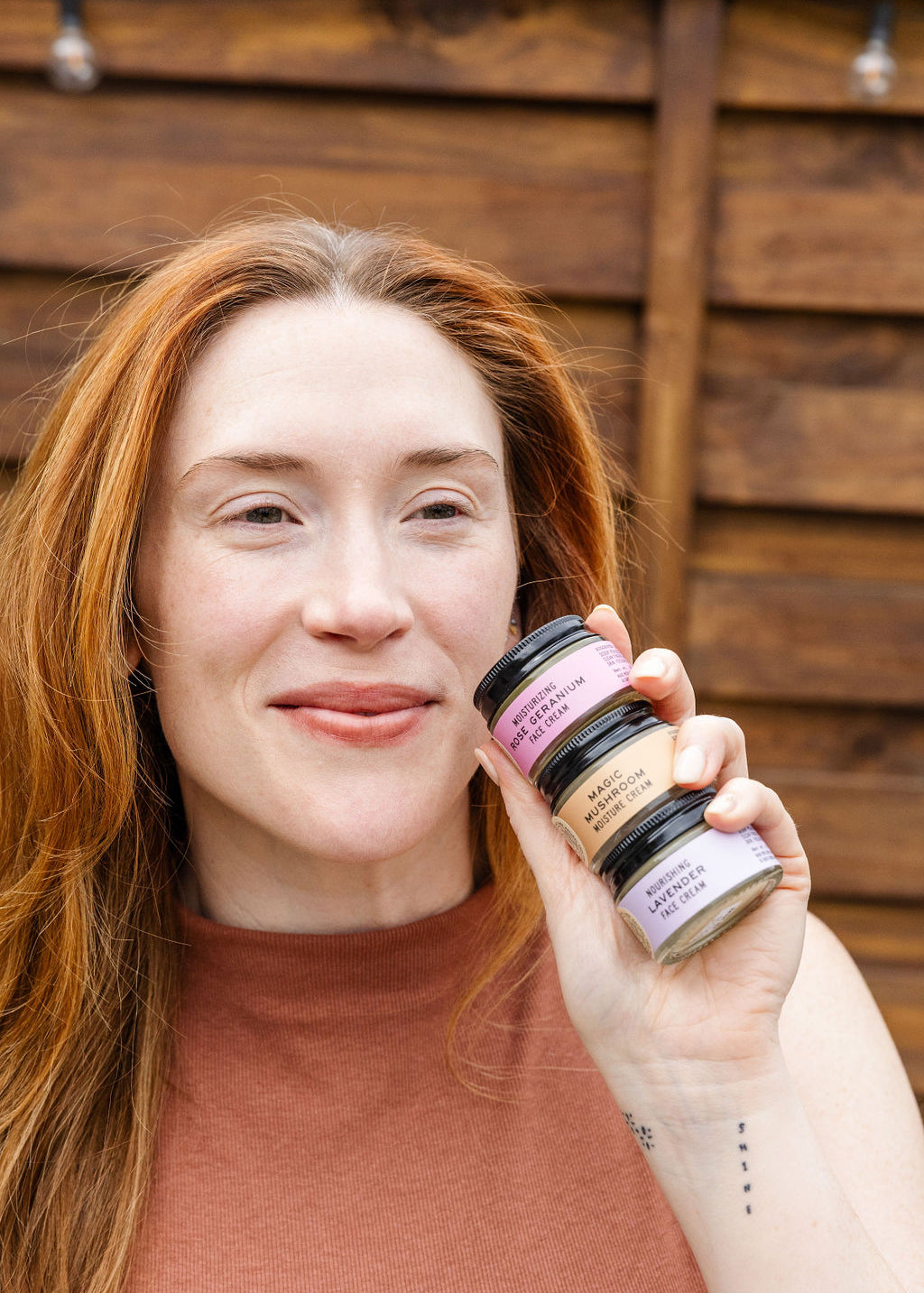 A woman with long red hair holds three stacked jars from the Good Flower Farm Facial Four Bundle near her face, smiling in front of a wooden background.