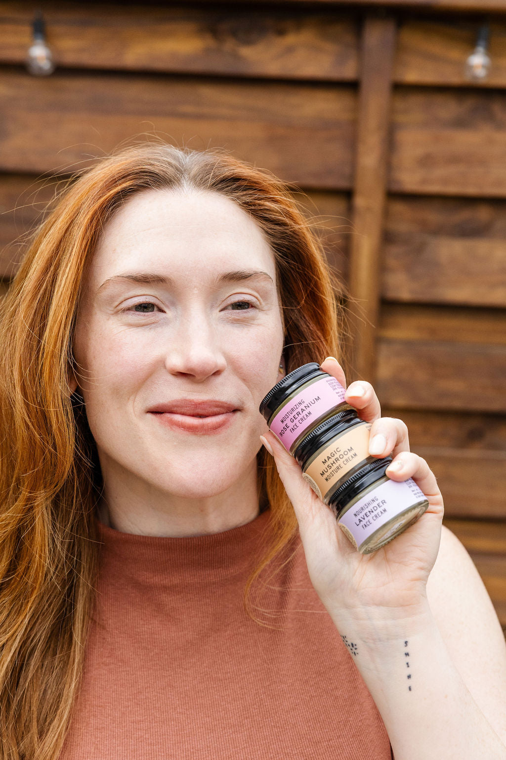 A woman with long red hair holds three stacked jars from the Good Flower Farm Facial Four Bundle near her face, smiling in front of a wooden background.