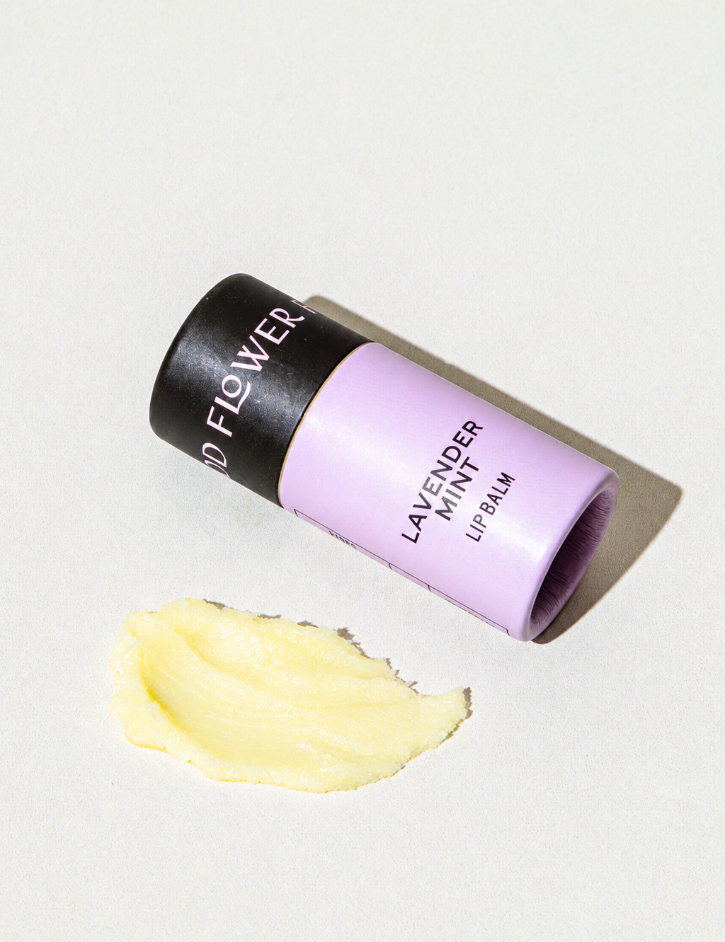 A lavender-colored cardboard tube of Good Flower Farm Lavender Mint Organic Lip Balm lying next to a smear of yellow balm.