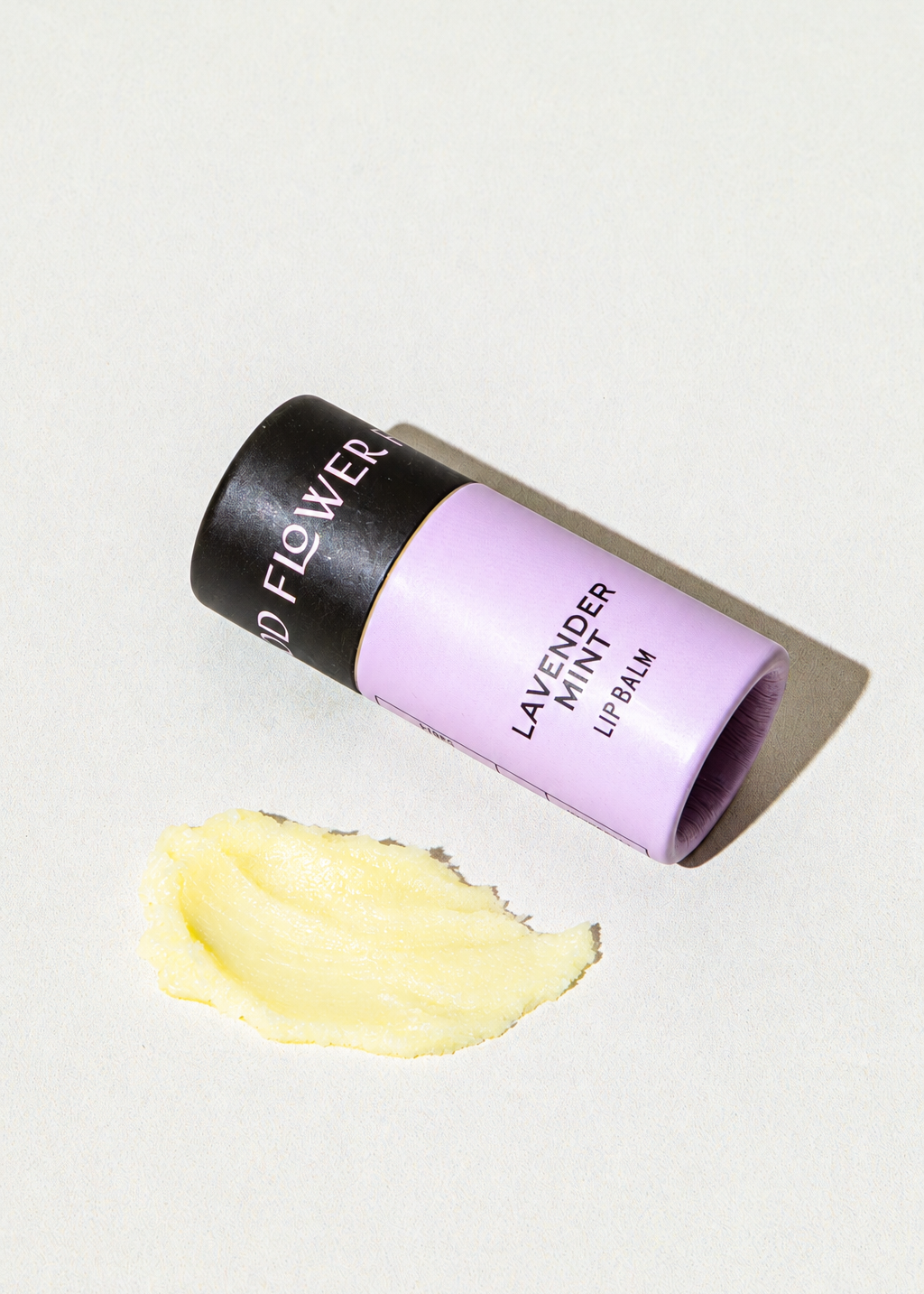 A lavender-colored cardboard tube of Good Flower Farm Lavender Mint Organic Lip Balm lying next to a smear of yellow balm.