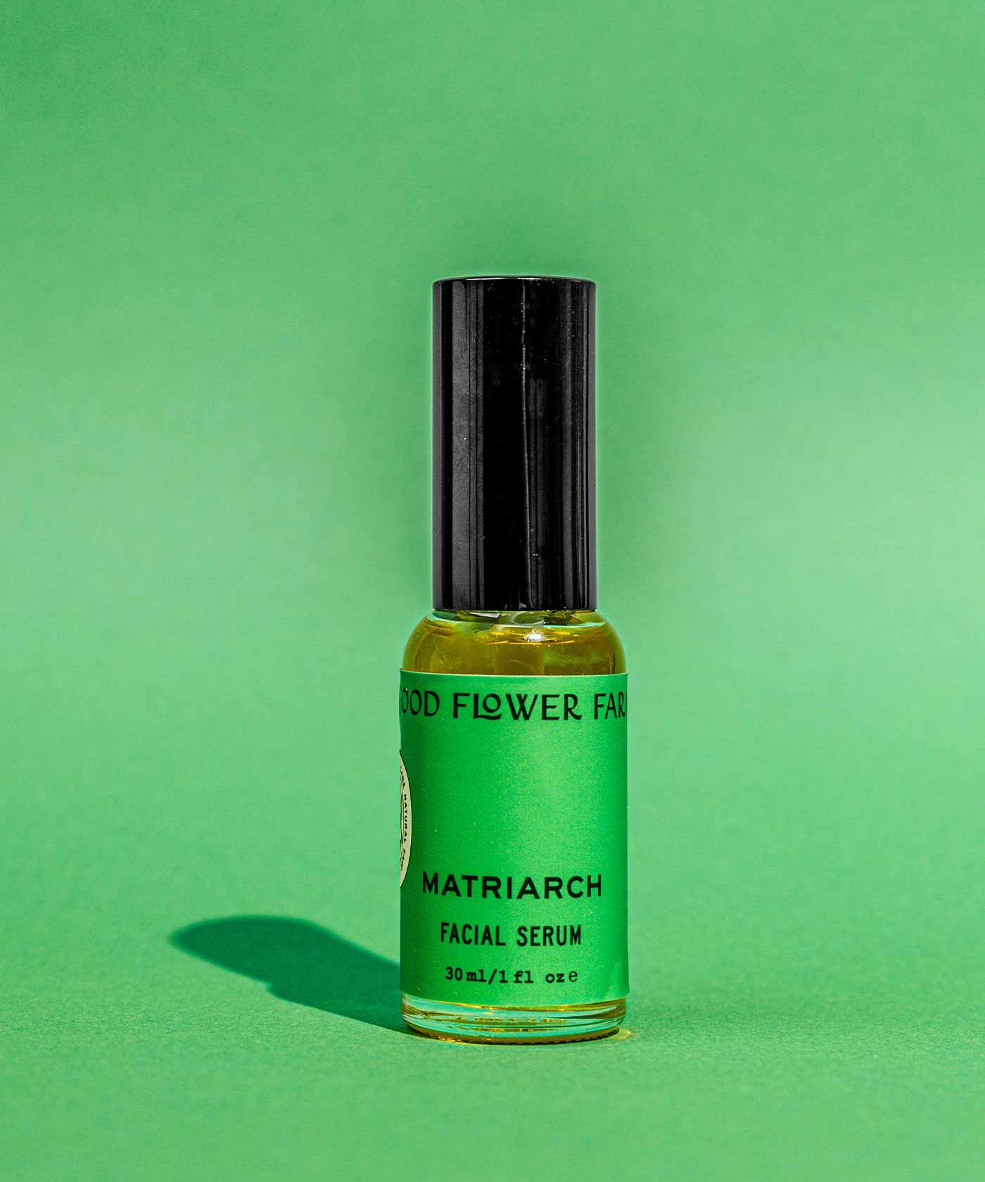 A small glass bottle of Good Flower Farm Matriarch Facial Serum, featuring a black cap and green label, stands upright on a green background. Infused with squalane, the serum casts a shadow on the surface.
