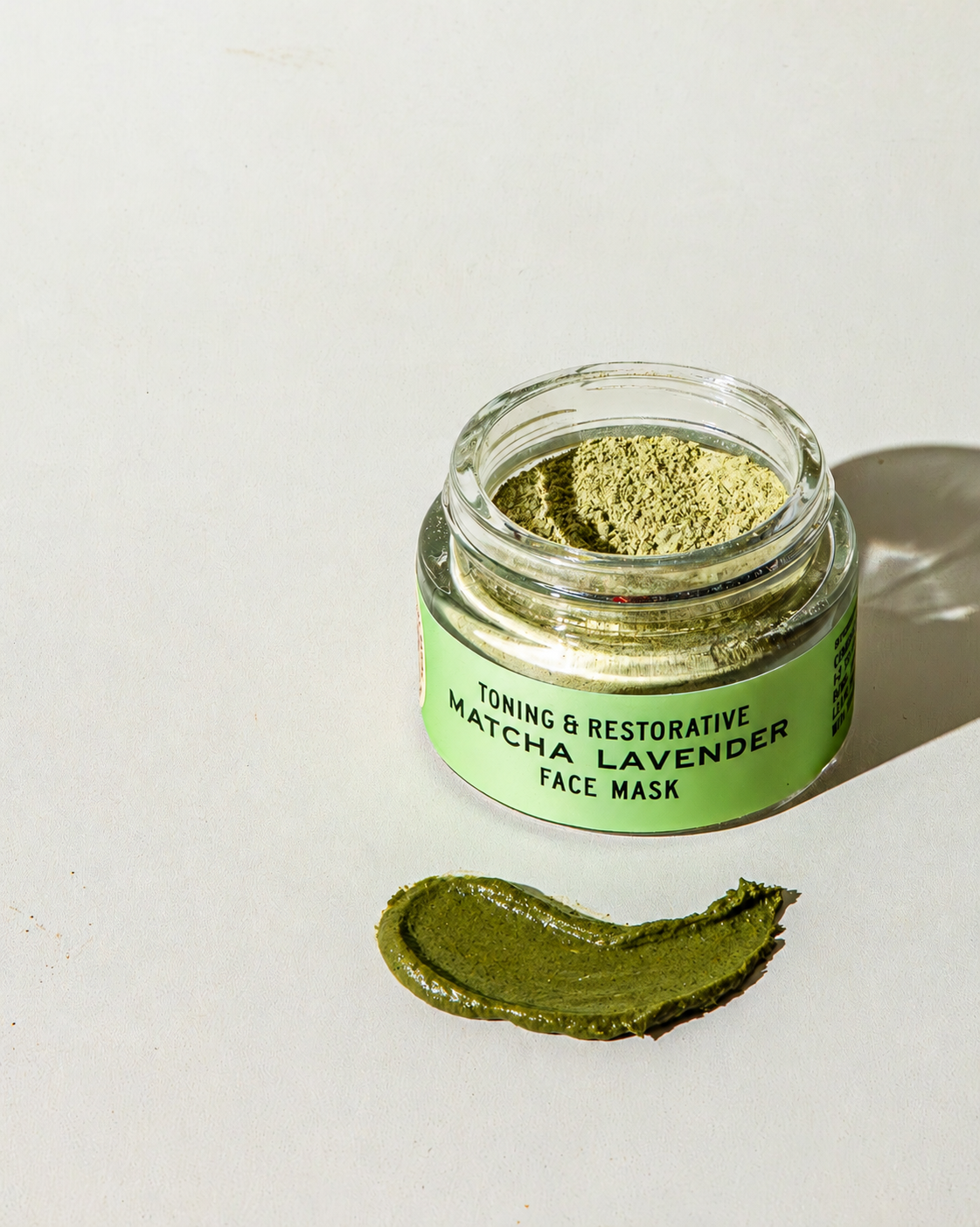An open glass jar of Good Flower Farm Matcha Lavender Botanical Face Mask filled with light green powder, with a smear of dark green paste in front of it.
