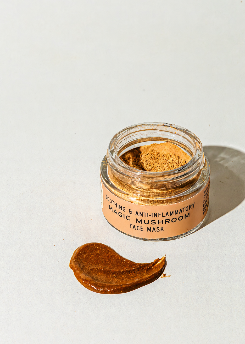 An open glass jar of Good Flower Farm Magic Mushroom Face Mask powder next to a smear of the product on a white surface, with a label reading Soothing & Anti-Inflammatory Magic Mushroom Face Mask.