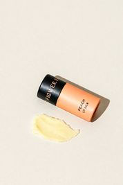 A peach-colored tube of Peach Organic Lip Balm by Good Flower Farm with a black cap, next to a small smear of deeply moisturizing yellow balm.
