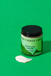 A jar of Woodland Dream Organic Body Cream with a black lid and a dollop of cream in front of it.
