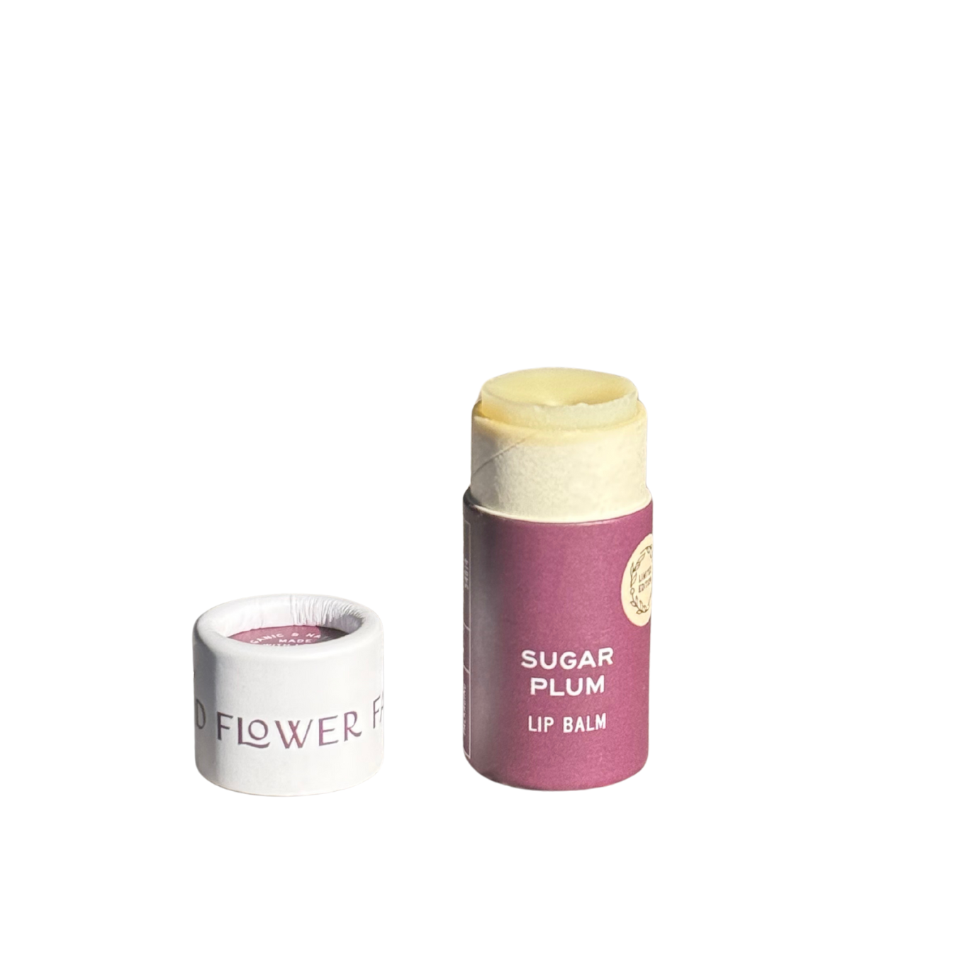 A tube of Good Flower Farm SEASONAL LIMITED Sugar Plum Organic Lip Balm stands upright with its cap off and beside it, revealing the moisturizing, eco-friendly balm inside. The lip balm features a purple Sugar Plum label and is set against a plain white background.
