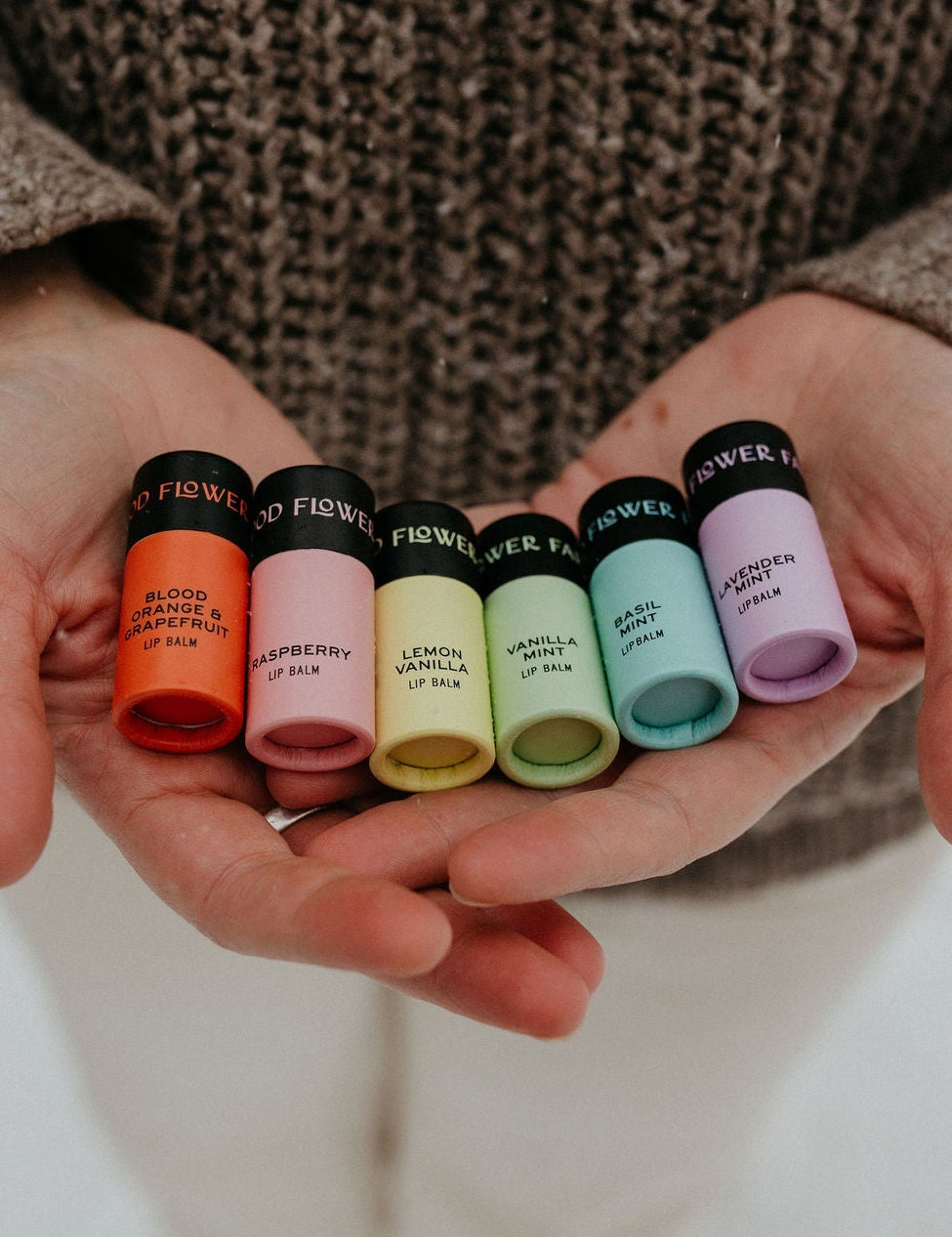 A person in a brown sweater holds six colorful Rainbow Organic Lip Balms by Good Flower Farm, arranged from red to purple, against a neutral background.