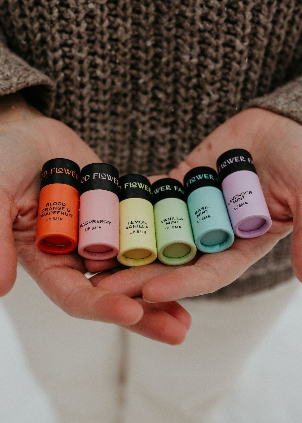 A person in a brown sweater holds six colorful Rainbow Organic Lip Balms by Good Flower Farm, arranged from red to purple, against a neutral background.