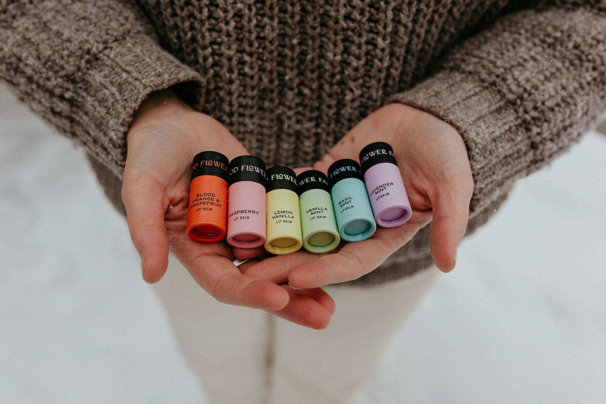 A person in a brown sweater holds six colorful Rainbow Organic Lip Balms by Good Flower Farm, arranged from red to purple, against a neutral background.