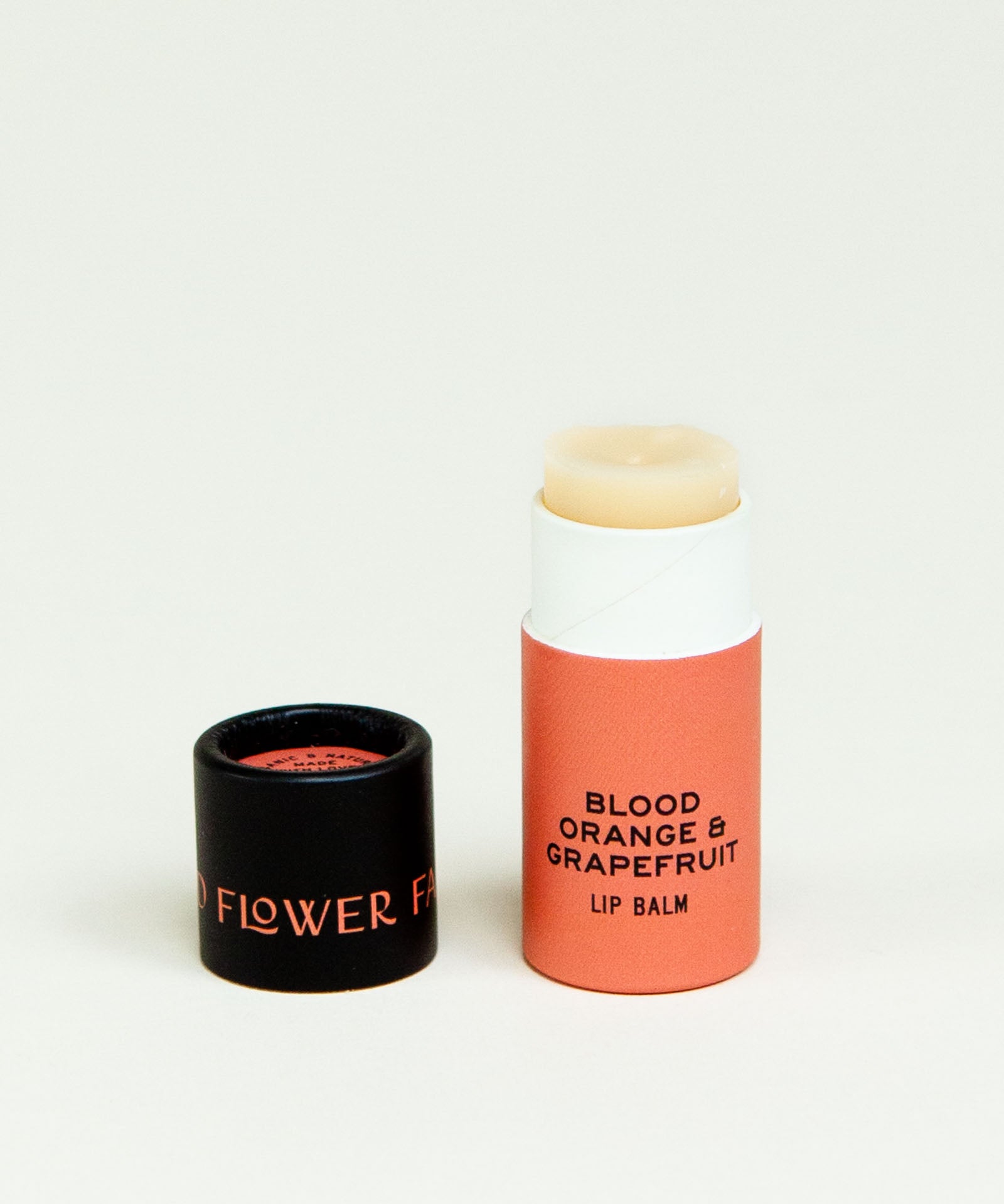 A tube of Seconds Lip Balm from Good Flower Farm stands upright with its black cap beside it on a plain white background.