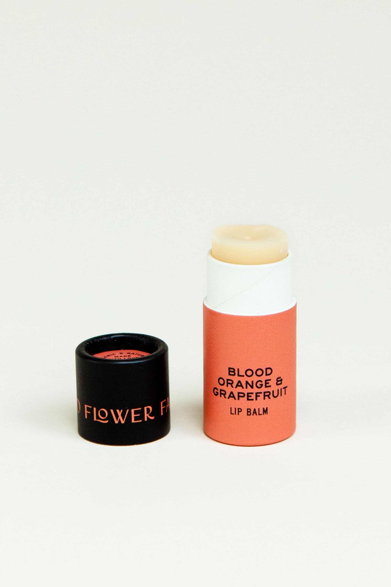 A tube of Seconds Lip Balm from Good Flower Farm stands upright with its black cap beside it on a plain white background.